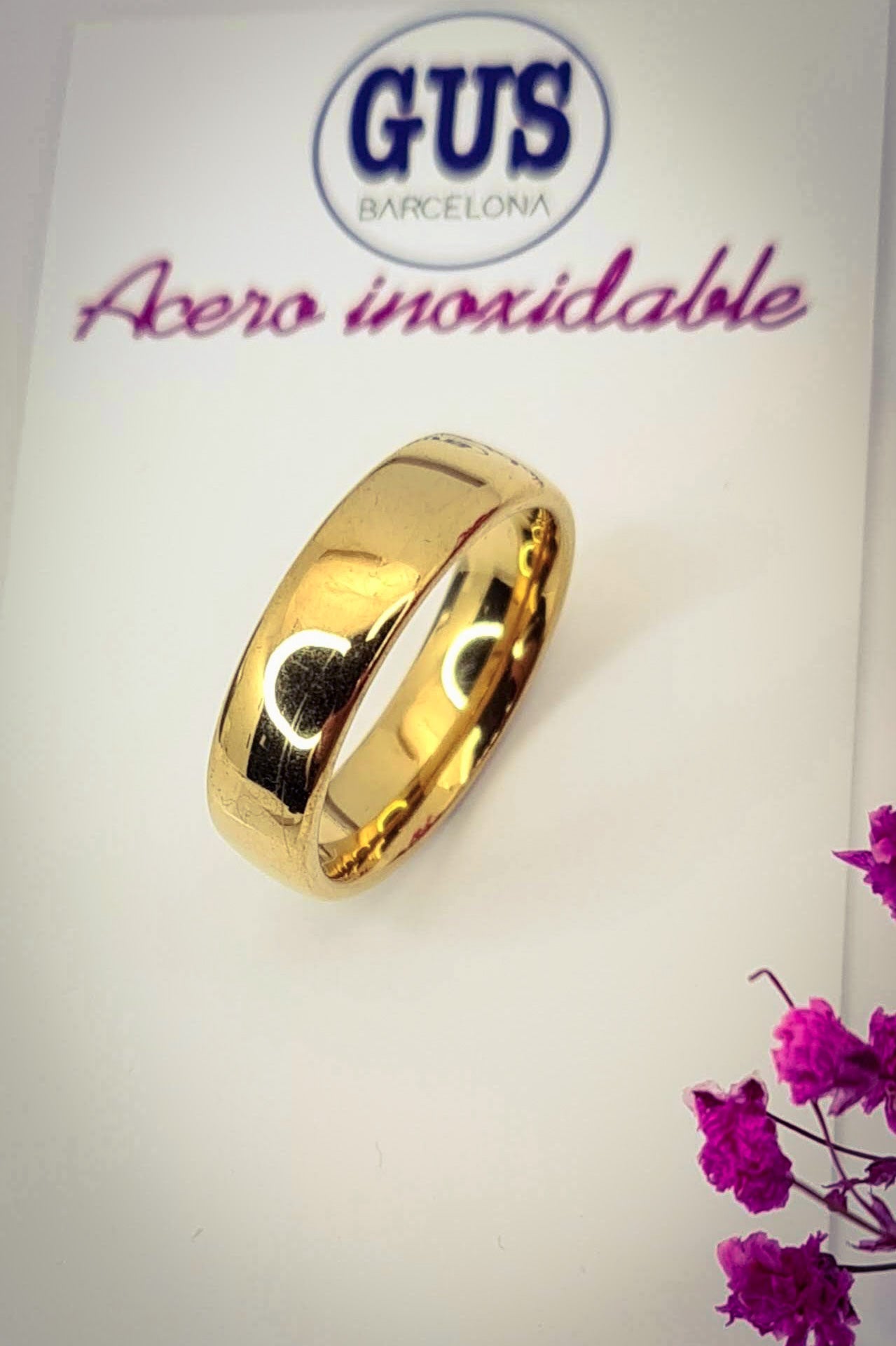 Anillo tipo alianza de matrimonio o pareja en acero inoxidable en color dorado y ancho de 6mm. El anillo está sobre la base de un cartón con el logo gus barcelona. Se incluye grabado de nombres y fecha importante gratis.